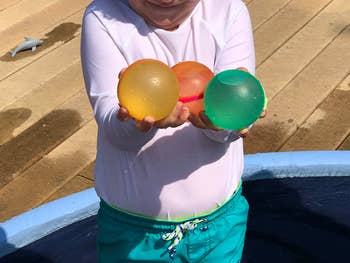 Child holding three water balloons by a kiddie pool on a sunny day