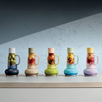 Five blenders on a counter, each filled with fruit and vegetables