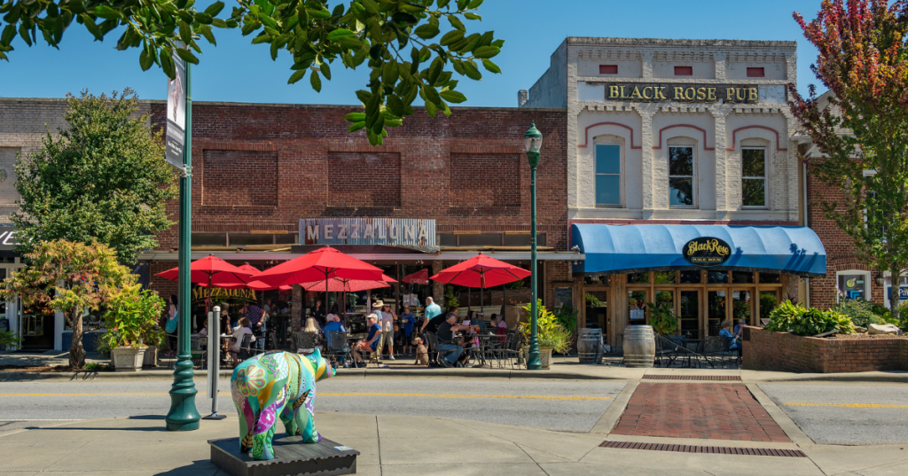 A street-level view of a vibrant, walkable downtown sidewalk in Hendersonville, lined with storefronts, outdoor seating, and shade trees.