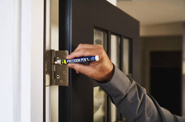 A hand uses a precision pen to lubricate a door hinge, demonstrating home maintenance