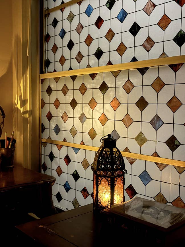 Stained glass window with geometric patterns casting warm light; a decorative lantern and books are on a wooden table nearby