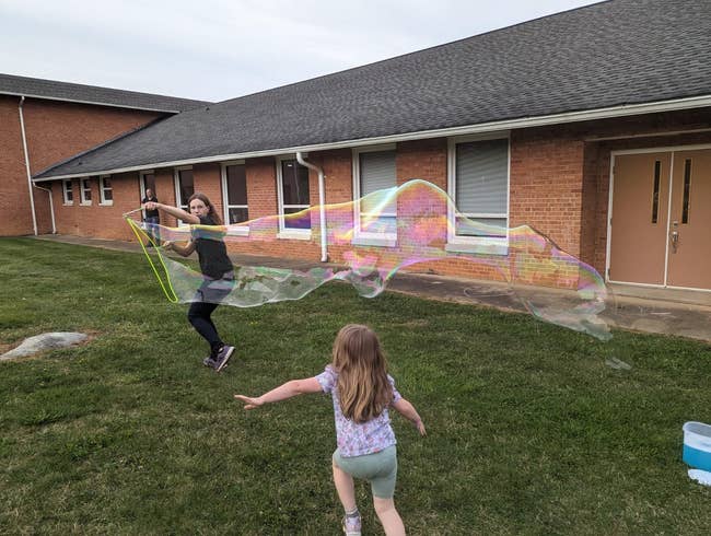 A person creates large soap bubbles with a wand outside a building as a child watches and runs toward them