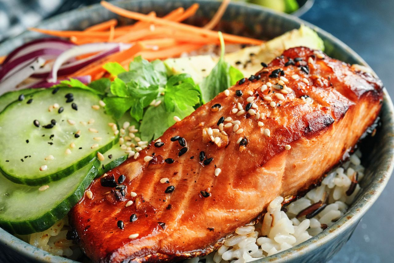 A plated dish featuring a piece of cooked salmon garnished with sesame seeds served over rice with sliced cucumbers carrots and herbs