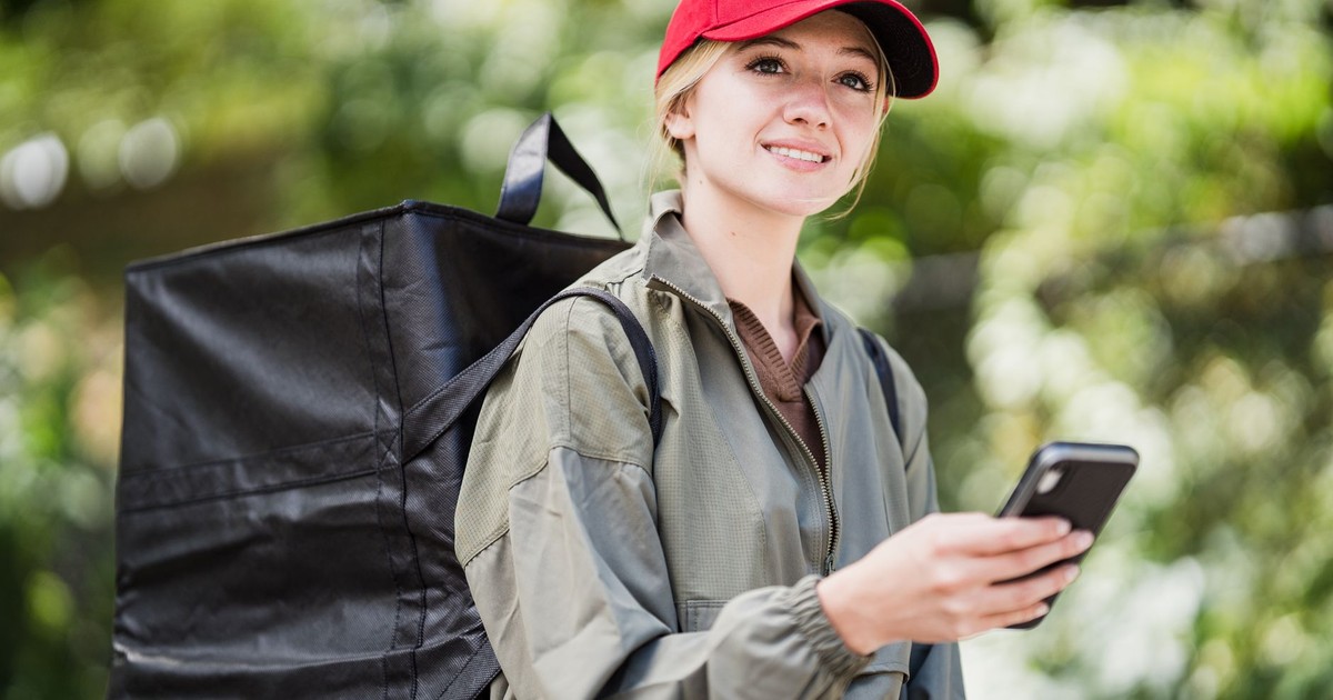 Female courier with smartphone using an app outdoors, wearing a red cap and green jacket.