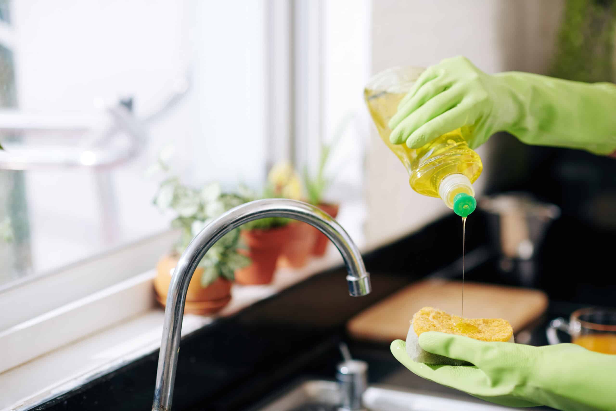 Closeup image of woman in latex gloves pouring dish soap on sponge and washing dishes
