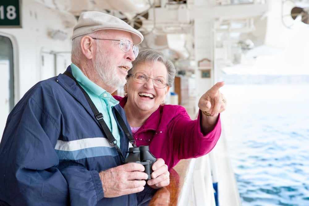 A happy elderly couple on a cruise ship, the woman points while the man holds binoculars, looking at the water.