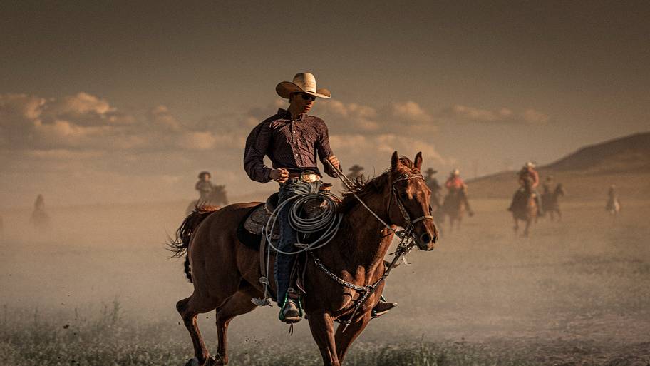 In foreground, a young cowboy on his horse during the run of the horses and, in background, a group of eight cowboys and cowgirls supervising the run of the horses.