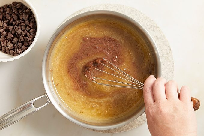 A hand uses a whisk to stir melted chocolate into a saucepan of brown batter, with a bowl of chocolate chips nearby on a white surface.