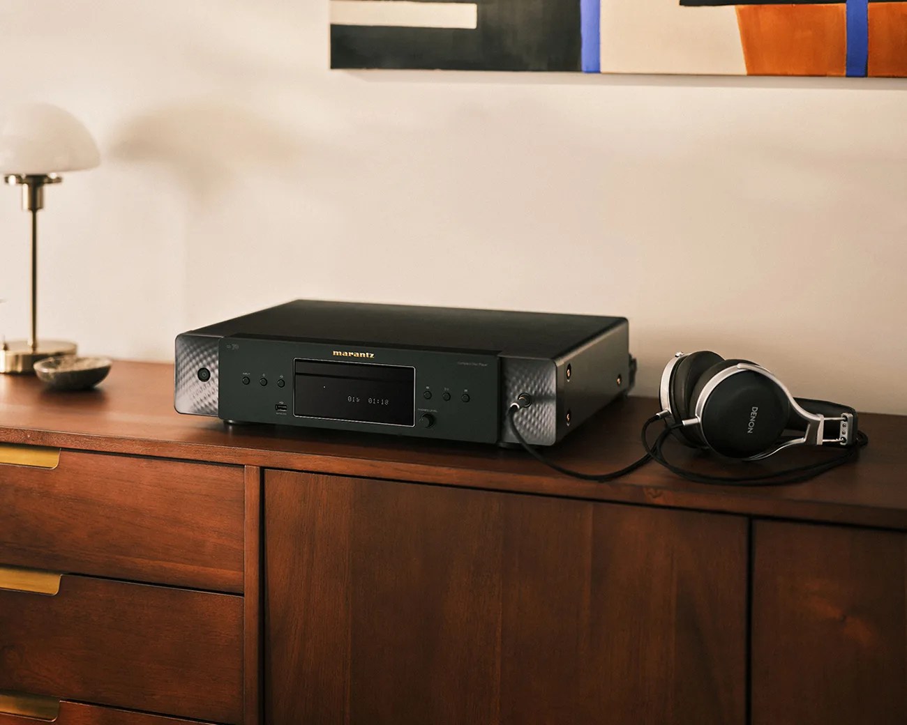 Black Marantz audio player with digital display and connected Denon headphones on a wooden cabinet.