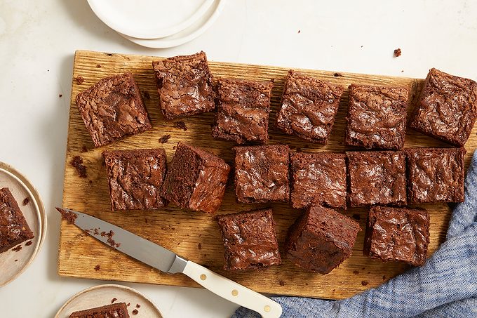 A wooden cutting board holds 20 square chocolate brownies, some with crumbly edges. A knife with a white handle rests beside them, and plates are partially visible nearby on a white surface.