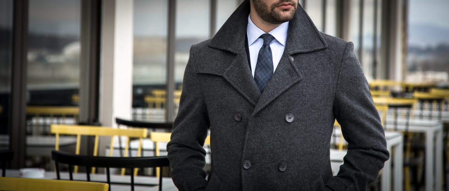 Man in tailored suit and trench coat standing and posing outdoors with his hands in his pockets