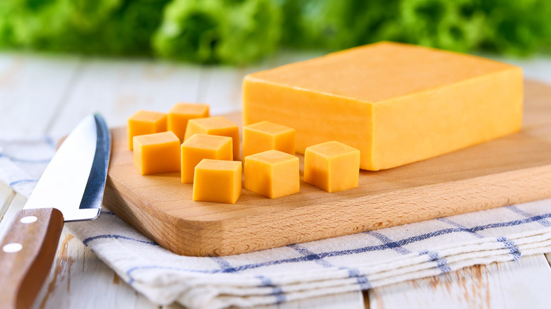 A block of cheddar cheese cut into chunks sits on a wooden cutting board.
