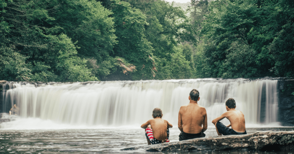 A wide view of Hooker Falls dropping into a calm swimming hole surrounded by dense green forest in DuPont State Recreational Forest. Three people sit in front of the falls.