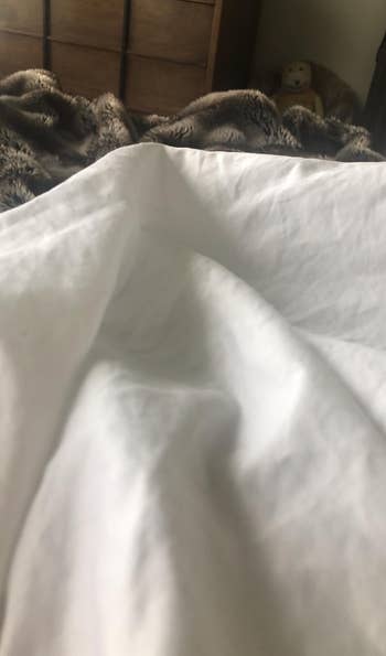 A close view of a white bedsheet with a blurred background featuring a teddy bear on a shelf