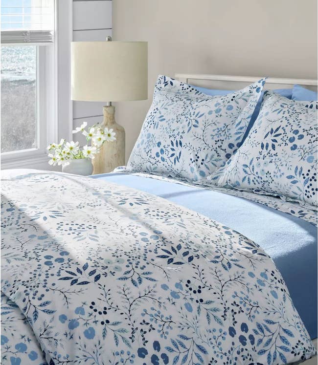 Floral bedding set on a neatly made bed, featuring a nature-inspired design, with a soft bedside lamp and flowers beside a window