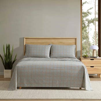 Gingham-patterned bedding set on a neatly made bed with a light wood frame, next to a nightstand with a lamp and a potted plant nearby