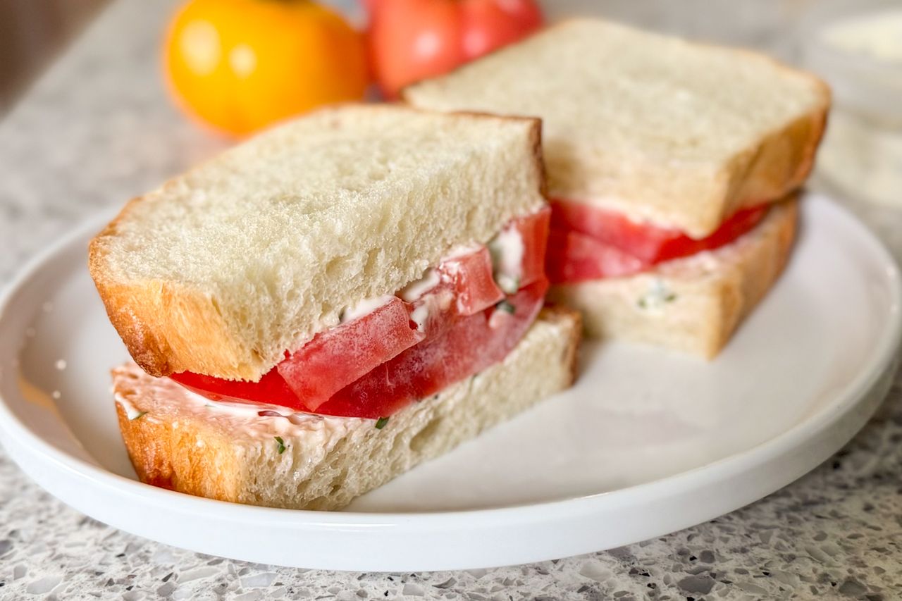 Two slices of bread with sliced tomatoes and a spread inside presented on a white plate with tomatoes in the background