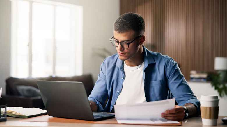 A person wearing spectacles and working on their laptop while holding a bunch of documents