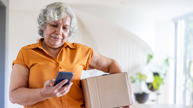 Mature woman holding a cardboard box while checking an app on her phone, as if looking up a shipping authorization for her package