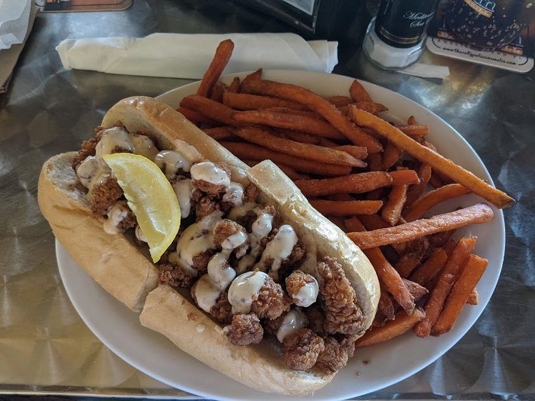 A po boy sandwich filled with fried seafood and creamy sauce is topped with a lemon wedge and served beside a large pile of sweet potato fries. The meal sits on a white plate on a metal table at The Salty Pelican in Fernandina Beach, Florida.