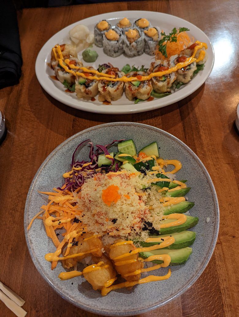 A large bowl holds a colorful sushi bowl with avocado cucumber shredded carrots purple cabbage crispy pieces roe and spicy sauce from Akari Sushi in Fernandina Beach, Florida. A second plate behind it contains two rows of sushi rolls topped with orange sauce and served with ginger and wasabi.