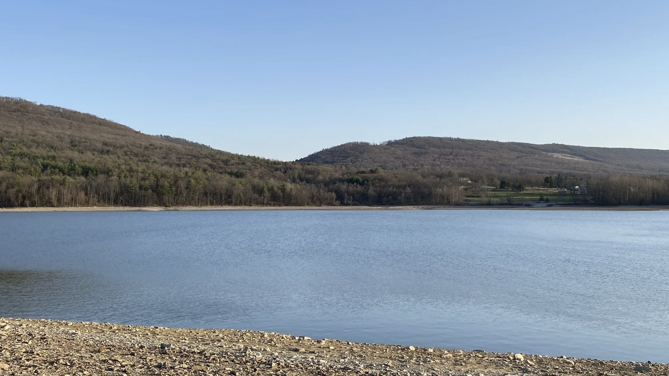 A rocky beach leads to a calm lake with mountains in the background.