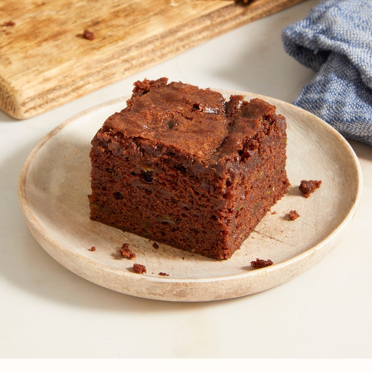 A single square piece of chocolate brownie sits on a round beige plate, with a wooden cutting board and a blue cloth visible in the background.
