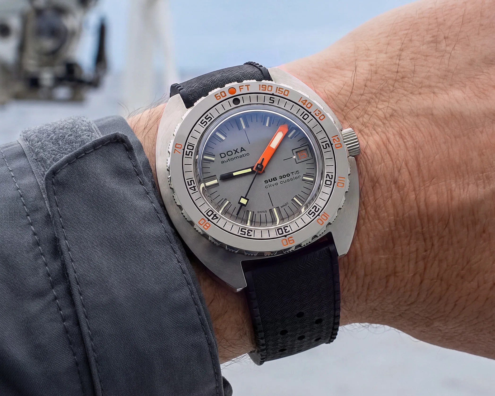 Silver DOXA SUB 300 Ti5 automatic wristwatch with gray dial, orange minute hand, and black perforated strap on a wrist.