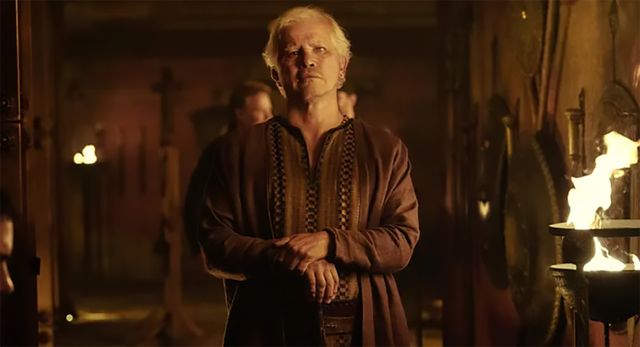 John Leguizamo in a medievalstyle scene standing indoors with a contemplative expression