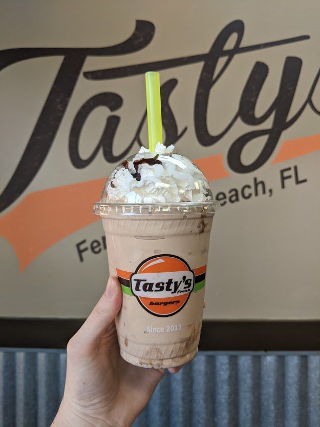 A hand holds a chocolate milkshake in a clear cup labeled