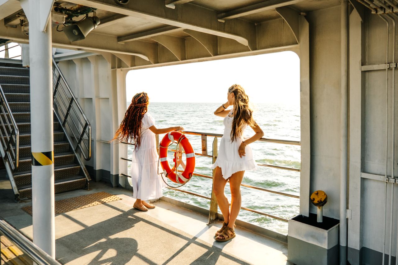 Two young women in white dresses standing on a ferry deck, looking out at the ocean.