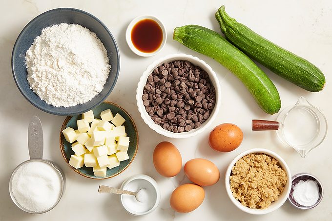 Baking ingredients including flour, sugar, baking soda, brown sugar, butter, eggs, chocolate chips, vanilla, milk, and two whole zucchinis are arranged on a countertop.