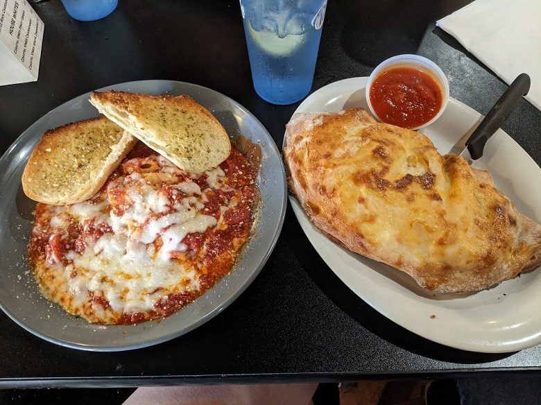 One plate holds a baked pasta dish covered in tomato sauce and melted cheese with two slices of garlic bread from OPS Pizza Kitchen & Café in Fernandina Beach, Florida. Beside it a large golden calzone is served with a cup of marinara sauce.