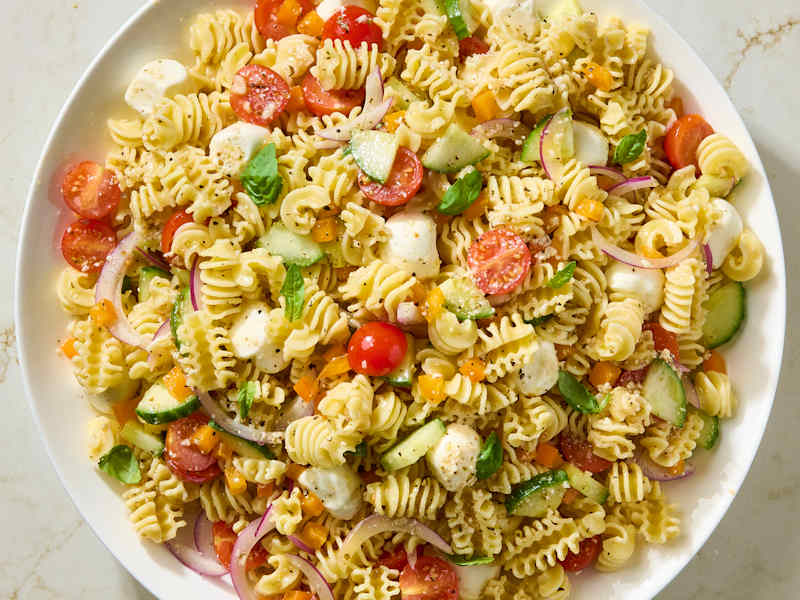 A large bowl of pasta salad with rotini, cherry tomatoes, cucumbers, mozzarella balls, red onion, and fresh basil.