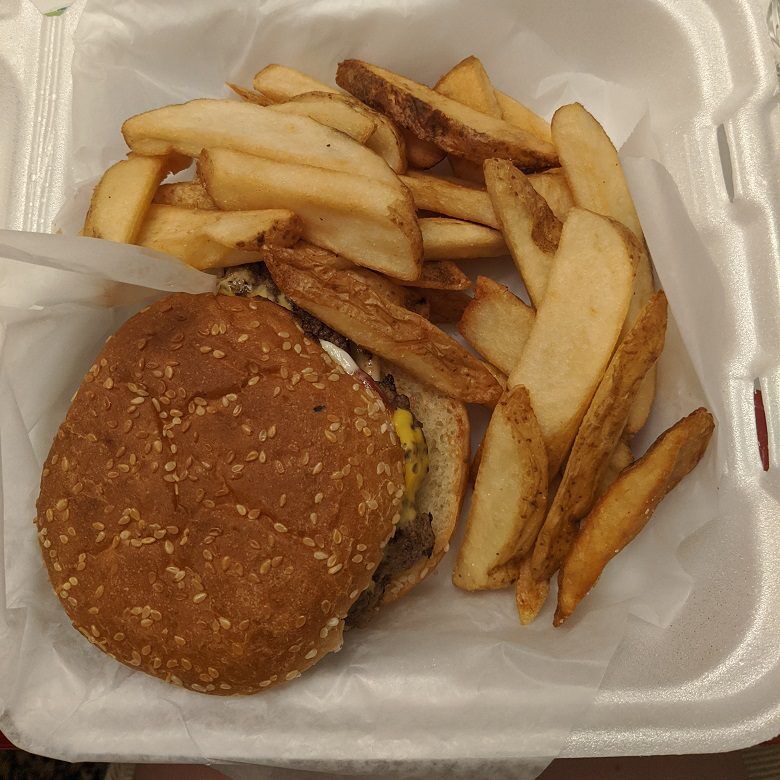 A cheeseburger on a sesame seed bun is served with thick cut potato wedges in a white foam takeout container from T-Ray's Burger Station in Fernandina Beach, Florida. Melted cheese and the beef patty are visible beneath the bun.