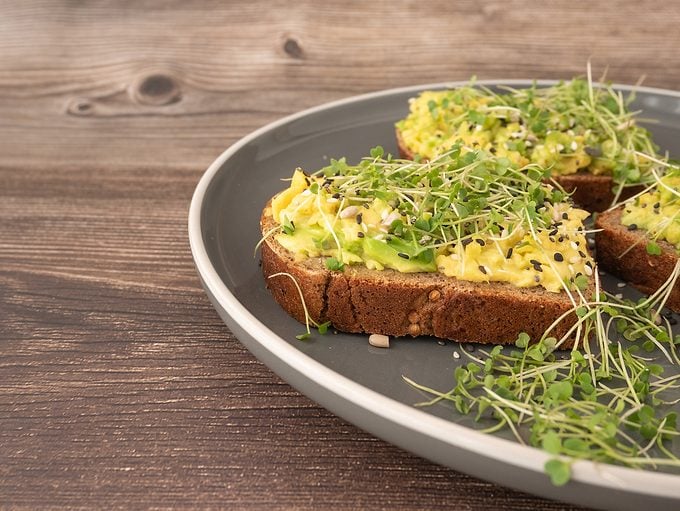 Micro Green Arugula Sprouts On An Avocado Sandwich On A Gray Plate On A Wooden Background. The Concept Of Proper Nutrition, Low Calorie Snacks And Weight Loss
