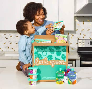 Parent and child happily open a Little Spoon box, revealing packaged children's meals on a kitchen counter, suggesting convenient, healthy meal options