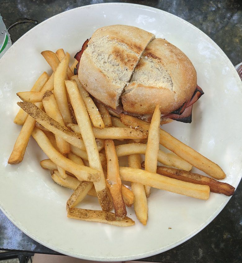 A toasted sandwich cut in half is served on a white plate with a pile of golden French fries from Cafe Karibo in Fernandina Beach, Florida. Slices of meat and melted cheese are visible inside the crusty round roll.