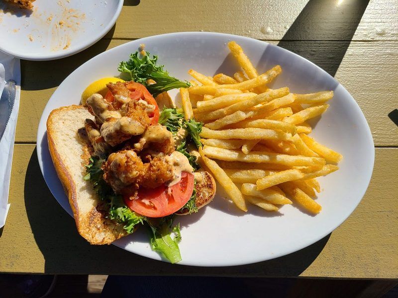 A fried seafood sandwich on a toasted roll is topped with lettuce tomato and creamy sauce and served with a large pile of French fries from Down Under Restaurant in Fernandina Beach, Florida. The meal sits on a white plate on an outdoor table in bright sunlight.