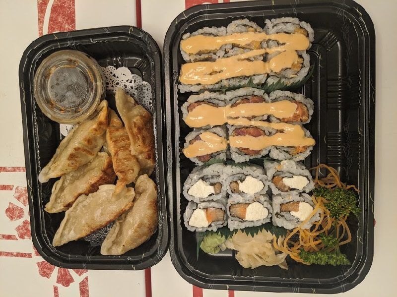 Two black takeout trays hold fried dumplings with dipping sauce and assorted sushi rolls topped with orange sauce from Hana Sushi in Fernandina Beach, Florida. Pickled ginger wasabi shredded vegetables and parsley garnish fill the remaining space.