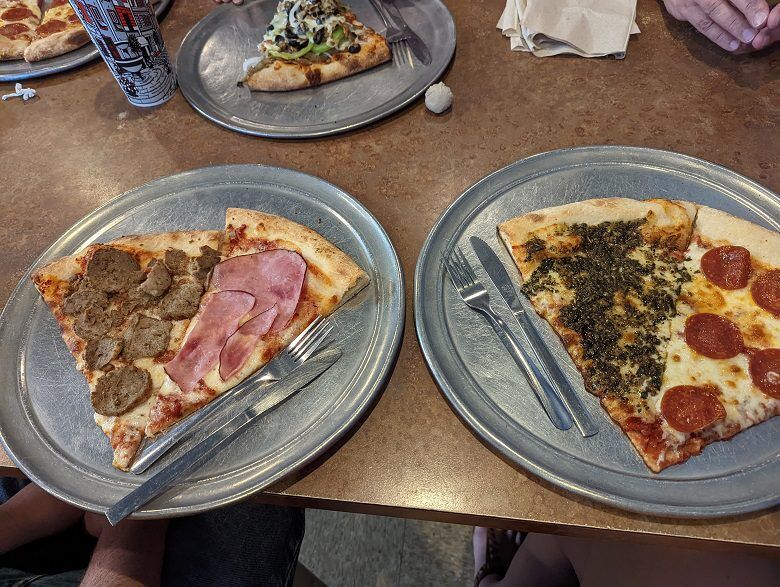 Two large pizza slices sit on round metal trays at Moon River Pizza in Fernandina Beach, Florida. One is topped with sausage and ham. The other is split between pepperoni and a dark herb covered topping.