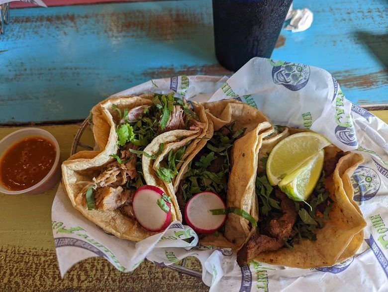 Three tacos filled with chopped meat and shredded greens are topped with radish slices and served with lime wedges and a side of red salsa. The tacos sit in a lined basket on a colorful wooden table at Cantina Louie in Fernandina Beach, Florida.