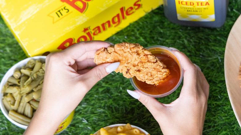 Someone dunks a Bojangles chicken tender into sauce, with iced tea and green beans in the background.