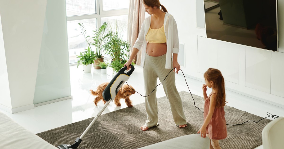 Pregnant mother cleaning carpet with vacuum as daughter and dog play indoors.
