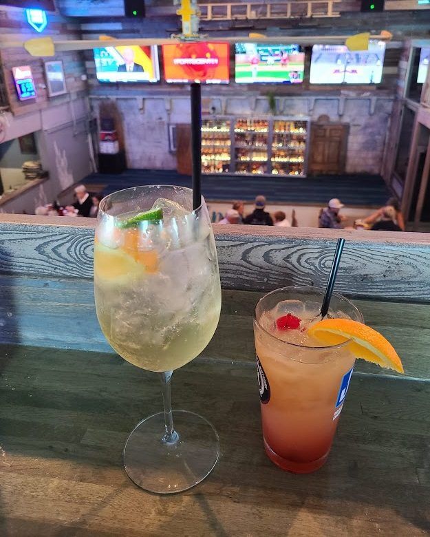 Two cocktails sit on a wooden ledge overlooking a busy sports bar with multiple televisions and rows of bottles at The Sandbar & Kitchen in Fernandina Beach, Florida. One drink is pale with lime and orange slices and the other is pink with an orange wedge and cherry.