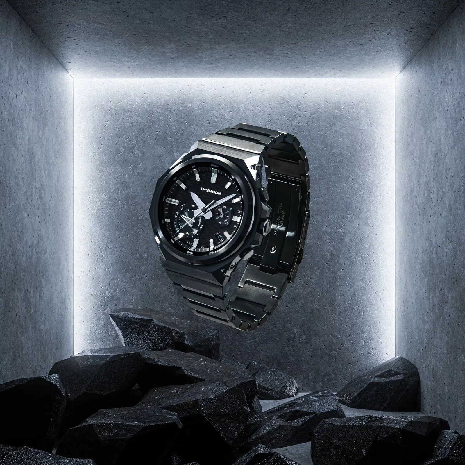 Black G-Shock stainless steel wristwatch with analog display floating above black rocks in a lit concrete corner.