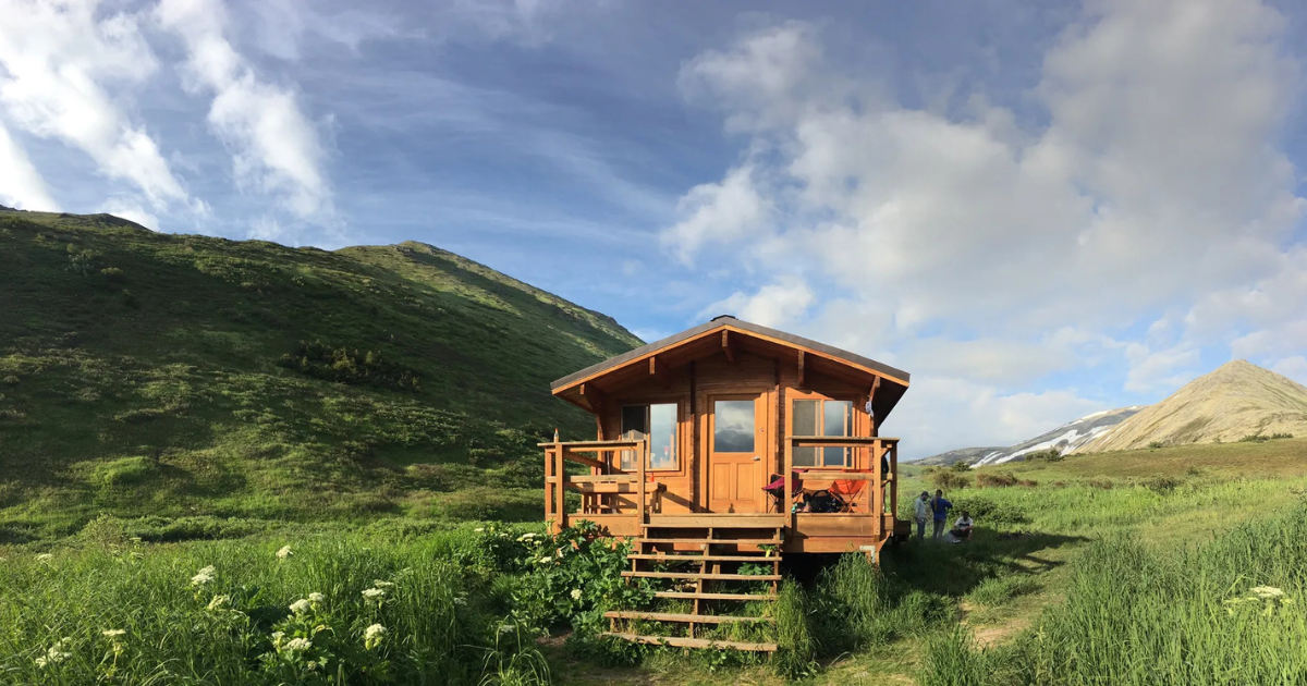 A cozy backcountry cabin sits in an alpine meadow along Devil's Creek Trail in Alaska, surrounded by rolling green hills and rugged snow-dusted mountains.