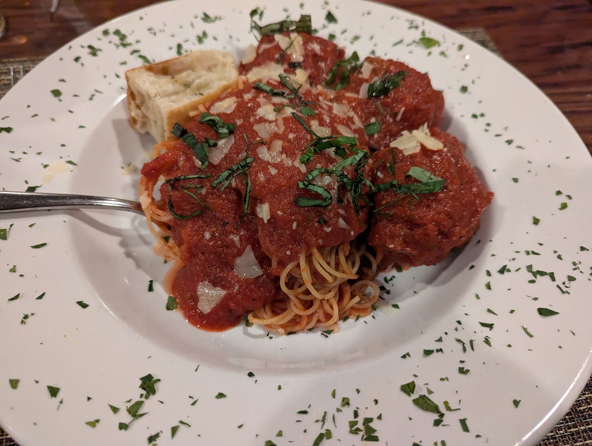 Spaghetti in tomato sauce is served with several large meatballs and topped with shaved cheese and fresh basil from Cucina South in Fernandina Beach, Florida. A piece of bread rests at the edge of the white plate.