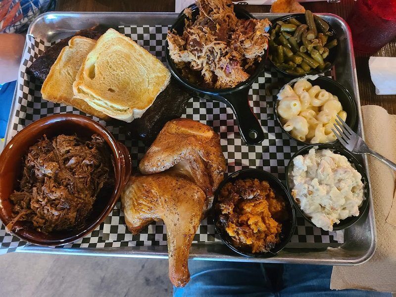 Barbecue platter with pulled pork from The Back Porch Southern Eats in Fernandina Beach, Florida. Smoked sausage. Two chicken leg quarters. Toast. Green beans. Macaroni and cheese. Potato salad. And sweet potato casserole served in small bowls on a metal tray.