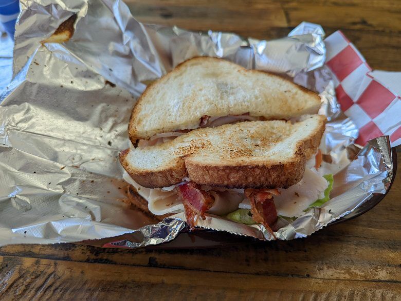 A toasted sandwich filled with sliced turkey bacon lettuce and tomato sits open on foil in a metal basket at 4th St. Deli & Salads in Fernandina Beach, Florida. A second sandwich half rests behind it on a wooden table.